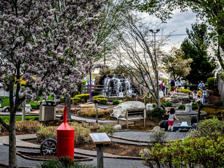 An outdoor scene depicts a park with people strolling, blooming trees, a red structure, pathways, rocks, and a waterfall in the background.