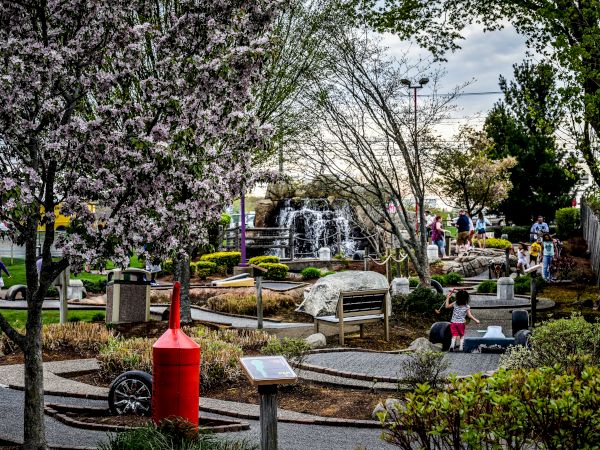 An outdoor scene depicts a park with people strolling, blooming trees, a red structure, pathways, rocks, and a waterfall in the background.
