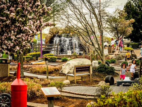 A park scene with children playing and adults walking near a fountain, surrounded by trees, benches, and greenery, and featuring a red buoy decoration.