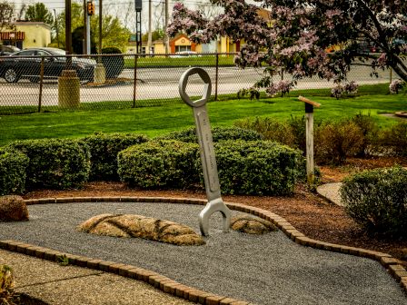 A large wrench sculpture is in a well-maintained garden near a street, surrounded by greenery and flowering trees.