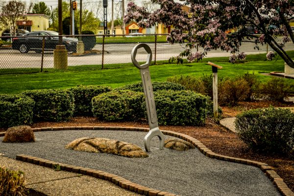 A large wrench sculpture is in a well-maintained garden near a street, surrounded by greenery and flowering trees.