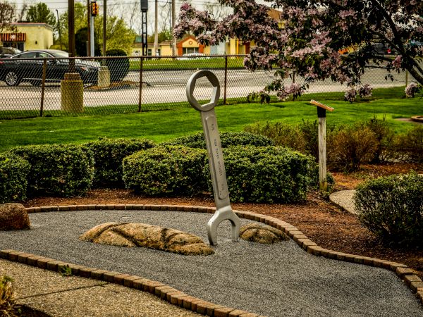 A large wrench sculpture is in a well-maintained garden near a street, surrounded by greenery and flowering trees.