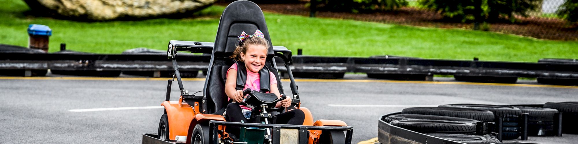 A child is driving a go-kart on a track with a large rock and greenery in the background. The child appears to be enjoying the ride, smiling.