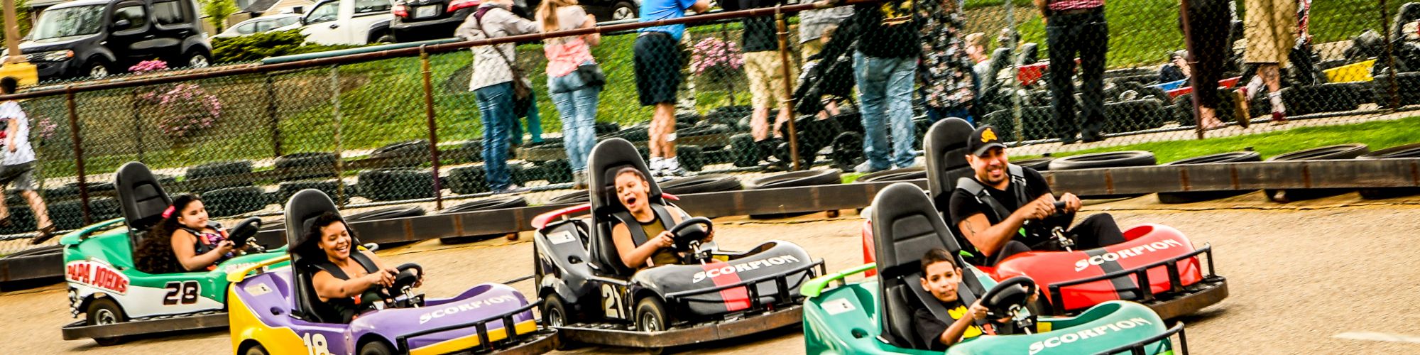 People driving go-karts on a track, with onlookers behind a fence.