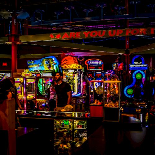 The image shows a vibrant, brightly lit arcade with various gaming machines and neon lights, with people interacting with the games.