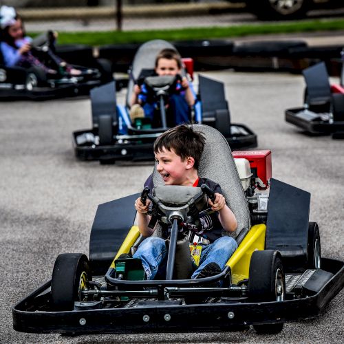 Children are driving go-karts on a track and appear to be enjoying themselves, with one child in the foreground looking ahead while smiling.