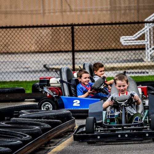 Three children are driving go-karts on a track lined with tires, with one child leading in a green kart followed by two in a blue one.