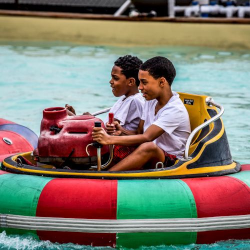 Two individuals are riding in a colorful bumper boat in a water amusement park, appearing to enjoy their time in the pool.