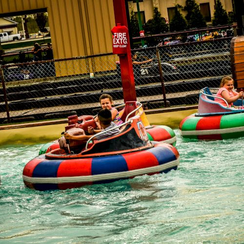 People are riding bumper boats on water, with colorful floating vehicles and a decorative pirate barrel in the background.