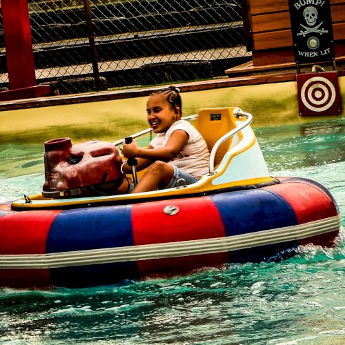 A young girl is enjoying a ride on a bumper boat in a water attraction, smiling and steering the boat, with a water target in the background.