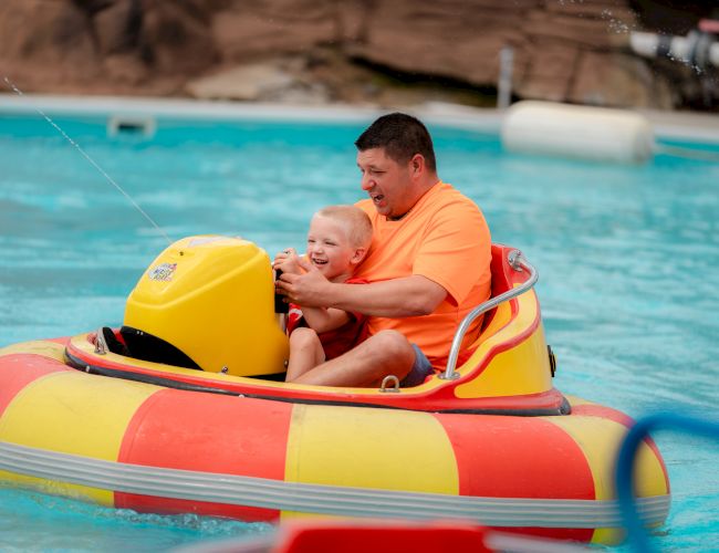 A man and child are enjoying a ride in a colorful bumper boat in a pool, smiling and having fun.