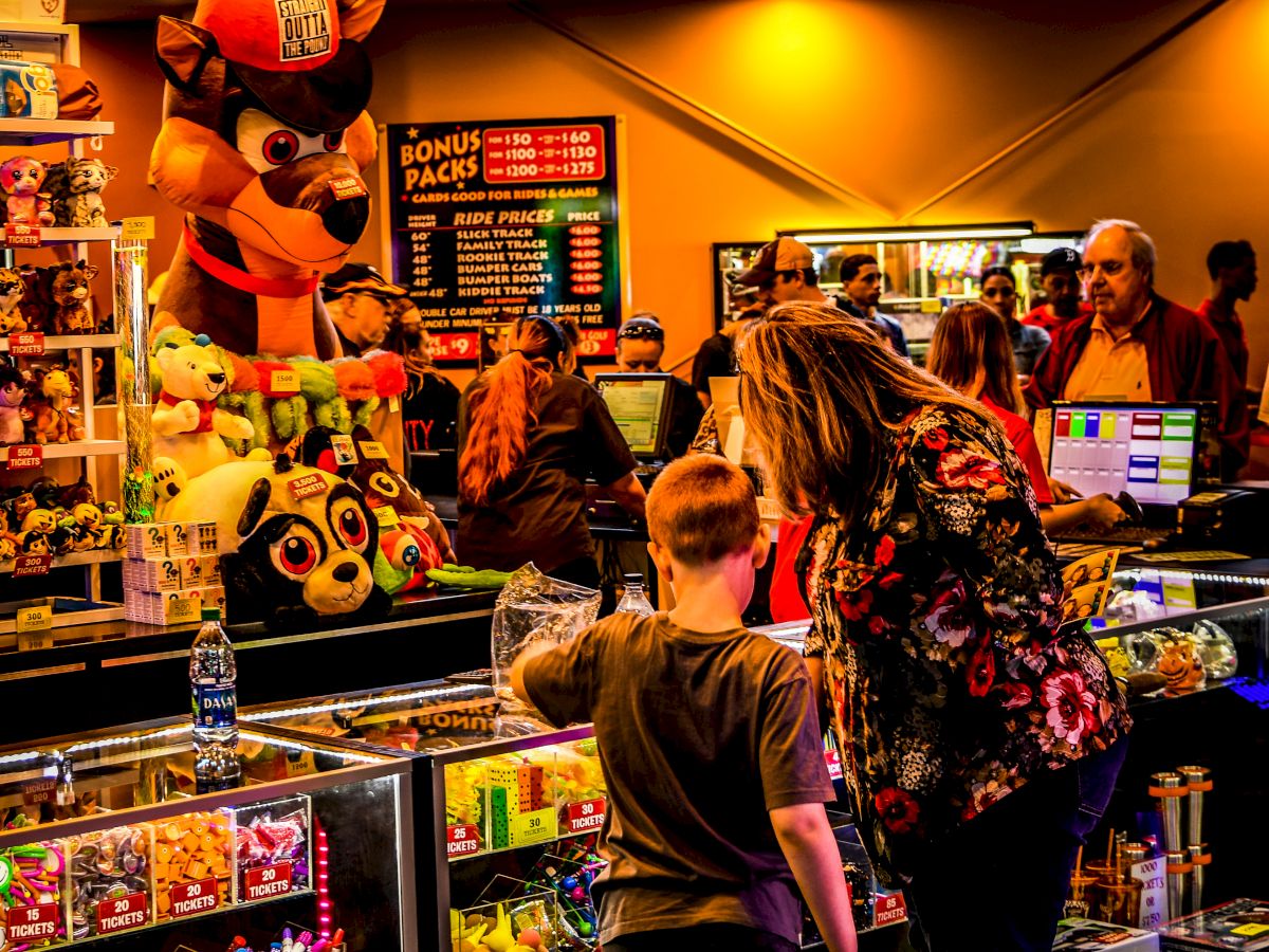 A lively arcade with several people interacting at the prize counter, displaying a variety of prizes and large stuffed toys in the background.