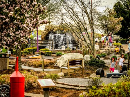 A park with pathways, a waterfall, benches, and various people, including children, exploring and enjoying the outdoor setting.