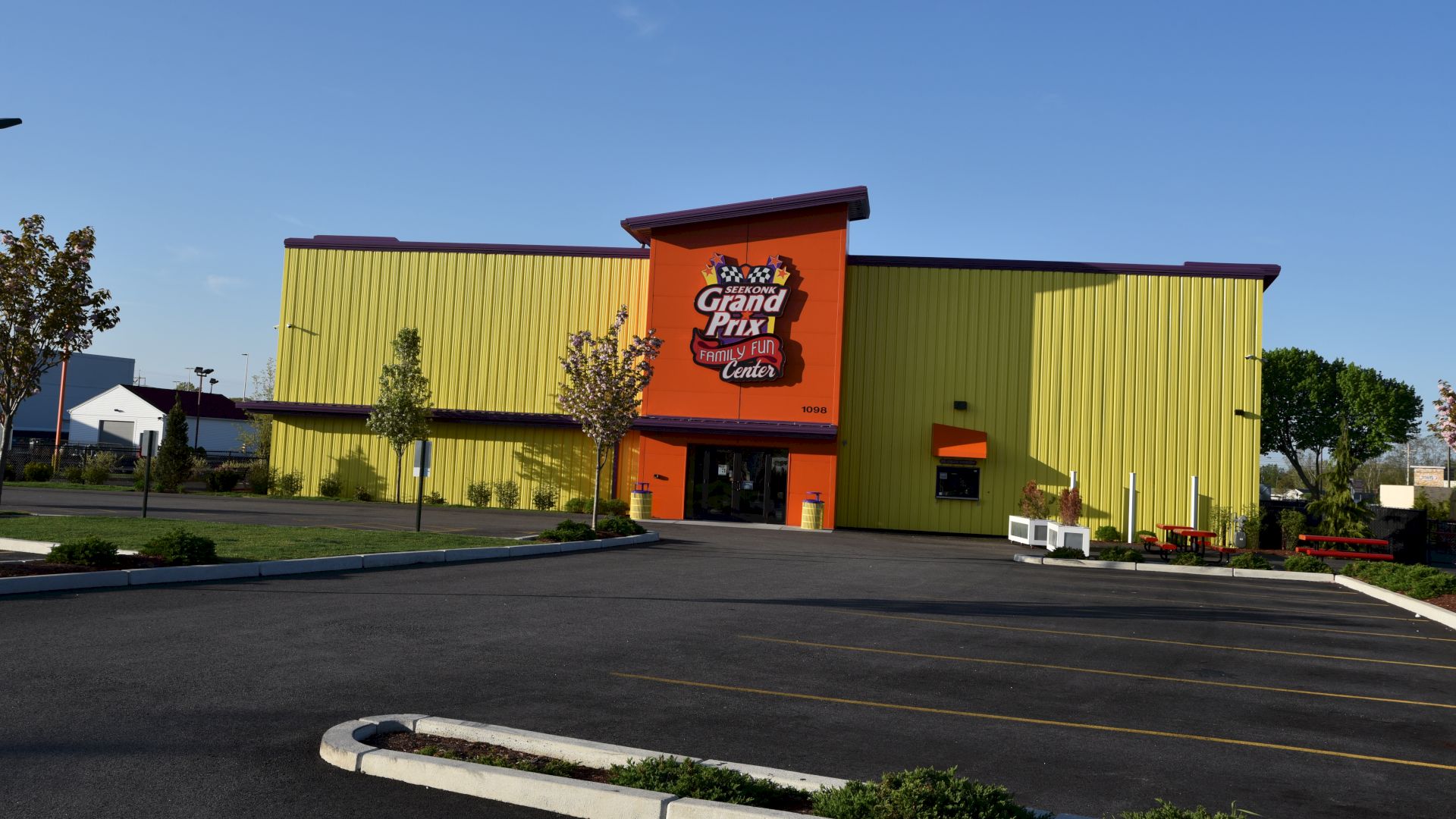 The image shows a brightly colored building with a "Grand Prix" sign, ample parking, and landscaped surroundings under a clear blue sky.
