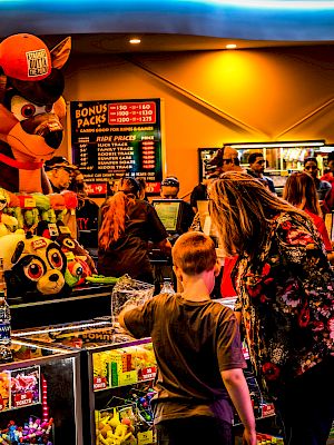 People are inside an arcade or amusement center, with a woman and child examining prizes. The area is filled with colorful games and lights.