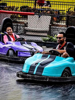 Two people enjoy a ride in colorful bumper cars, one blue with number 7, and one purple with number 10, at an amusement park.