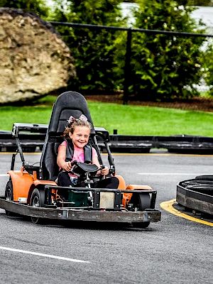 A child is driving a go-kart joyfully on a track with greenery and a large rock in the background, with barriers marking the track route.