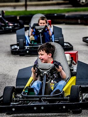 Children are driving go-karts on a paved track, appearing to enjoy the experience.