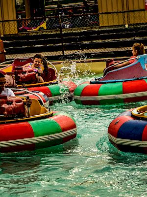 People are enjoying a bumper boat ride in a pool, splashing water and steering colorful boats, while having fun at an amusement park.