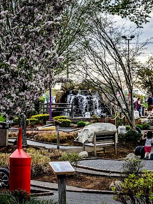 The image shows a park with blooming trees, walking paths, a red cylindrical sculpture, a small waterfall, and people enjoying the scenery.