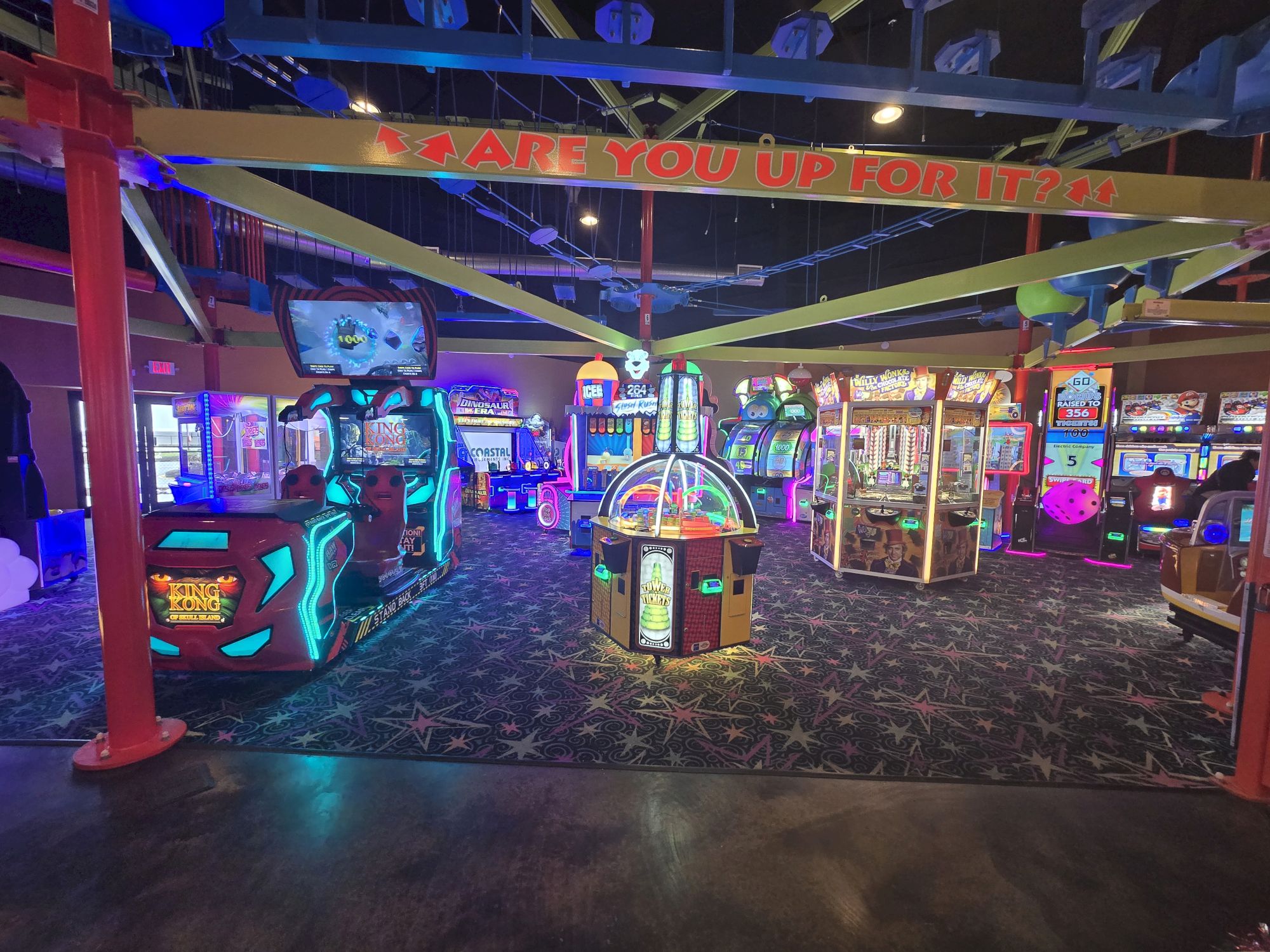 A vibrant arcade filled with glowing arcade games, neon lights, and a colorful central prize machine; futuristic machines line the dark, patterned floor, with banners overhead.