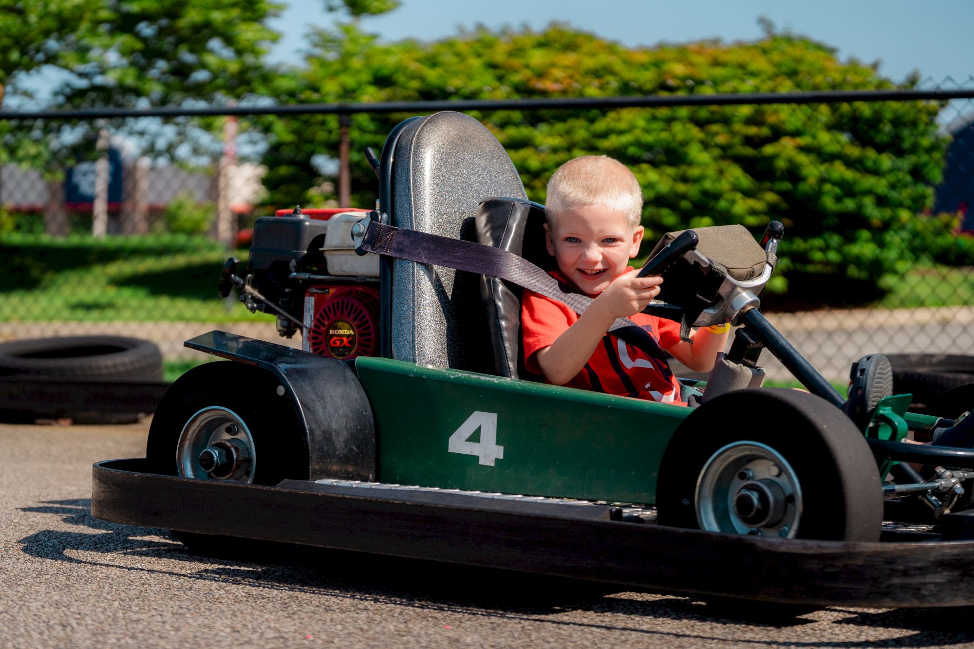 A child is happily driving a green number 4 go-kart on a track with a safety belt fastened, surrounded by a fence and greenery.