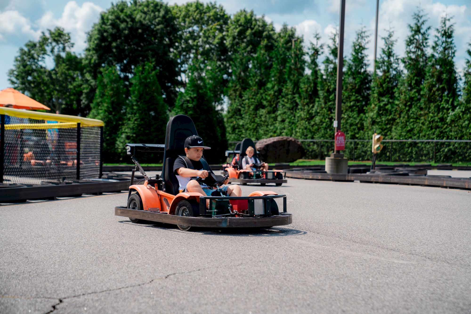 Two people are driving orange go-karts on an outdoor track surrounded by trees and barriers.