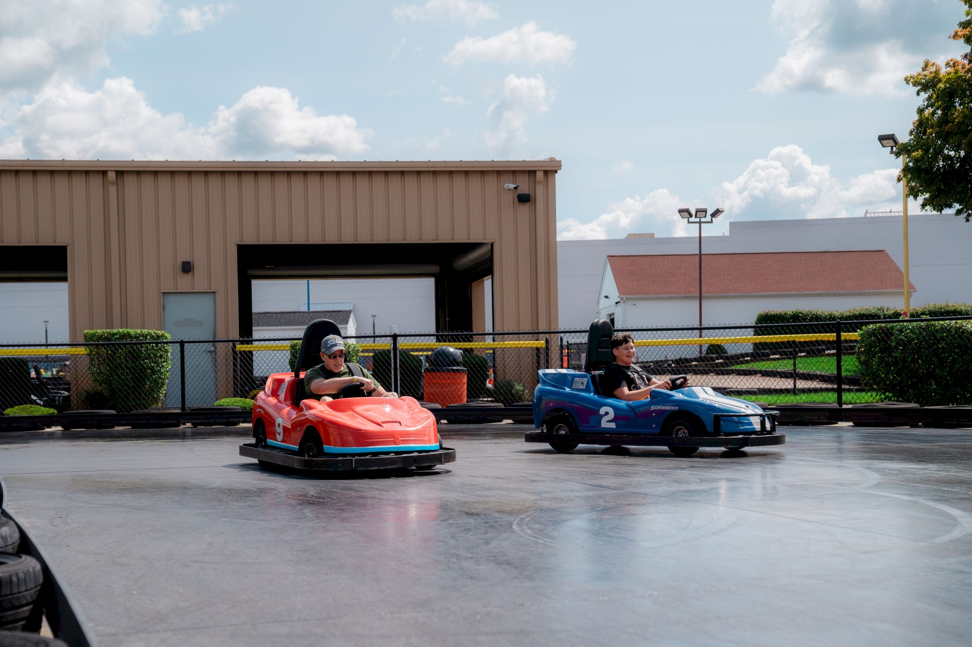 Two people are driving bumper cars on a track, with buildings and a fence in the background.