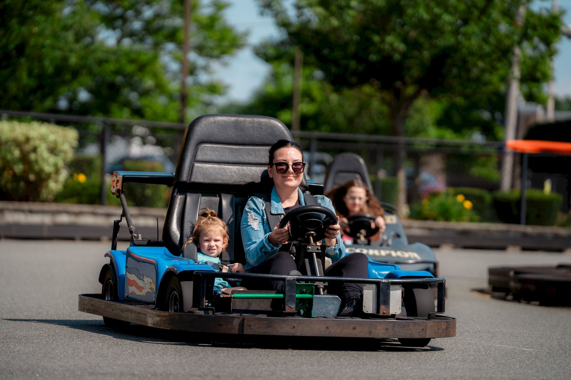 People driving go-karts on a track, with trees in the background. A woman and a child are in the front kart, enjoying the ride.