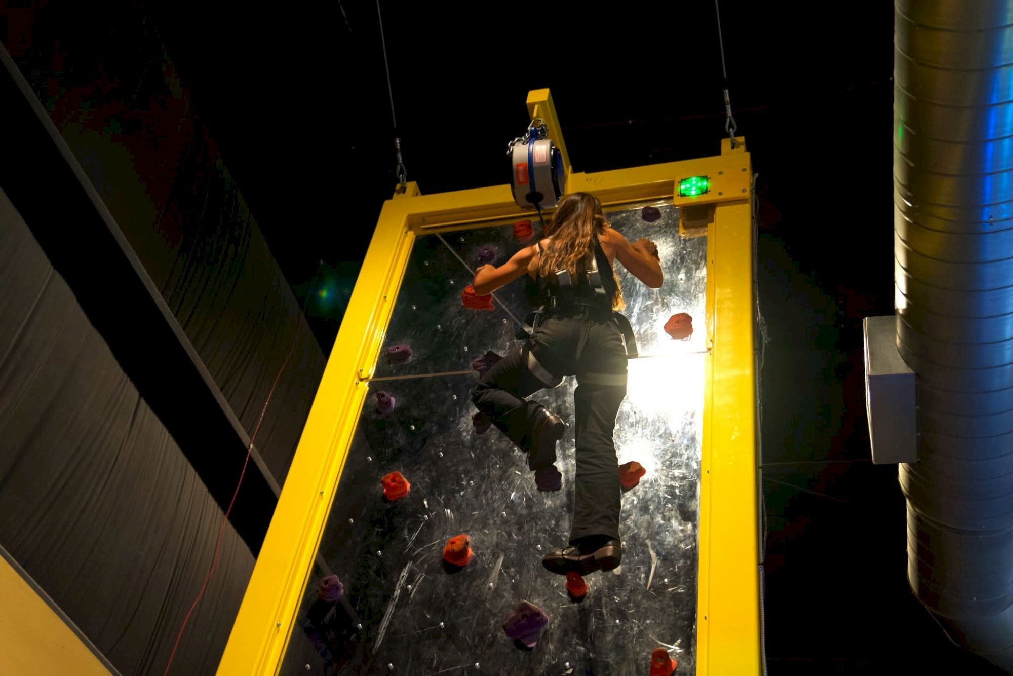 A person is suspended in a yellow climbing frame, mid-climb on an indoor rock wall with colorful holds, overhead lighting, and industrial surroundings.