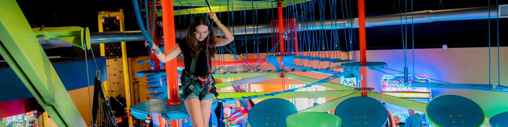 A vibrant indoor rope course with hanging obstacles, colorful ropes and nets, and people navigating high above in a themed arcade area.