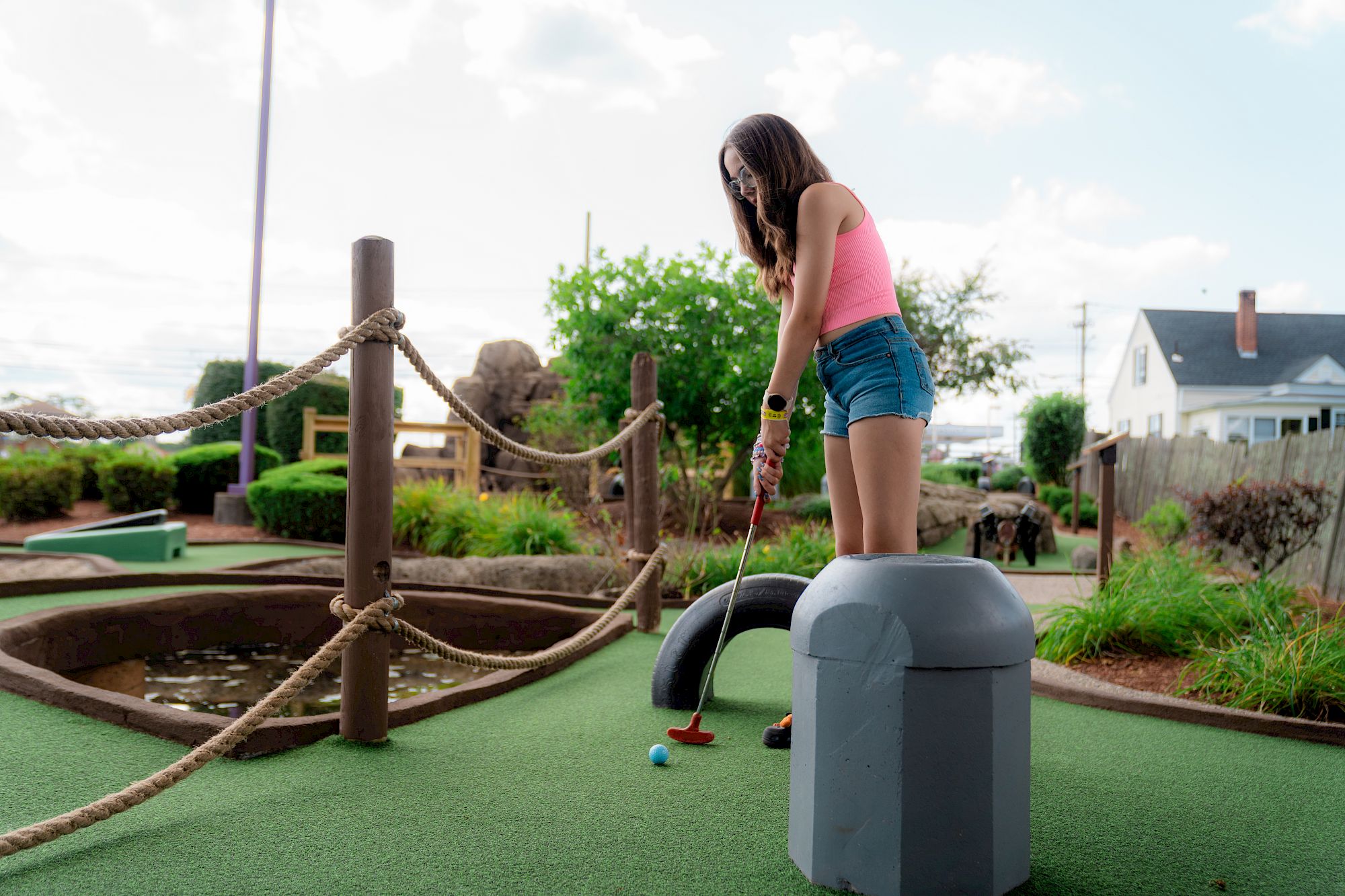 A person playing mini-golf, wearing a pink top and denim shorts, stands on artificial turf ready to putt a ball.