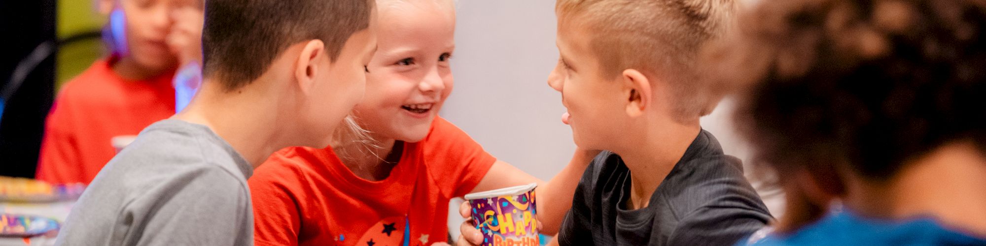 Kids are sitting at a table, smiling and holding party cups, with colorful decorations and snacks around them, enjoying a cheerful gathering.