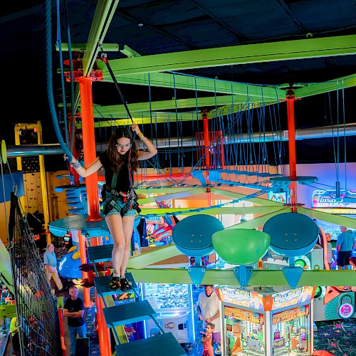 A person navigates a colorful indoor ropes course in an entertainment center. Bright lights and arcade games are visible below.