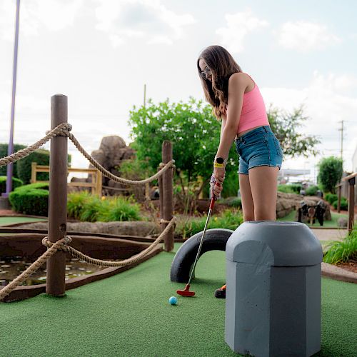 A person in casual summer attire plays mini-golf on a course with greenery and rope barriers under a clear sky.