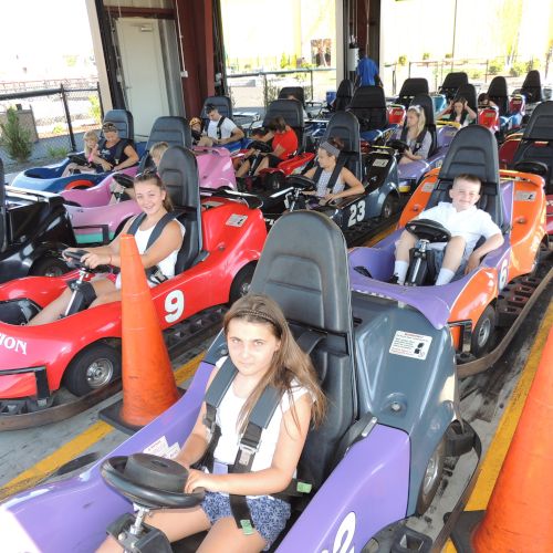 People sitting in go-karts, ready for a ride on a track. Orange cones are placed in front of some karts, and a building is visible in the background.