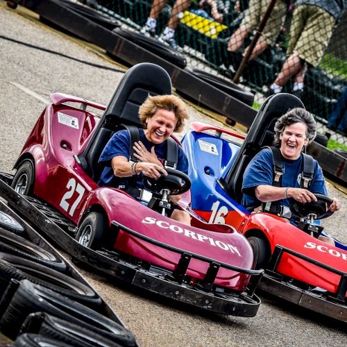 Two people are racing in go-karts on a track, wearing casual clothing and smiling. The go-karts are red and blue.