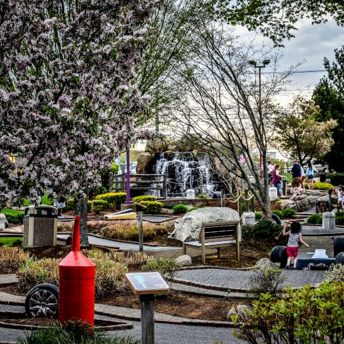 A serene park with blooming trees, pathways, a red structure, a waterfall, rocks, and people enjoying the surroundings can be seen.