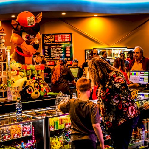 A bustling arcade with colorful prizes and large stuffed animals, showing a woman and child at the counter, while others enjoy the vibrant atmosphere.