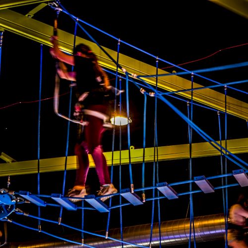 People are navigating a high ropes course indoors at night with brightly colored equipment and safety harnesses.