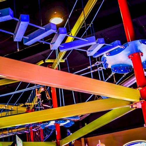 The image shows an indoor ropes course with colorful beams and platforms, featuring a participant navigating the elevated obstacles.