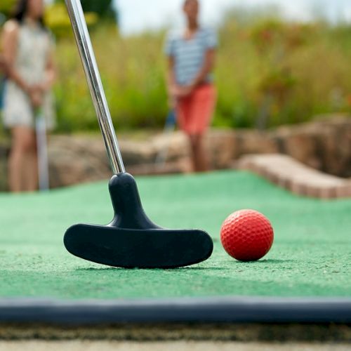 A close-up of a mini-golf putter and a red ball on a green surface, with two blurry, unidentifiable people in the background.