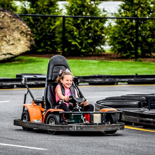 A child is driving a go-kart on an outdoor track, smiling and looking ahead. The track is surrounded by greenery and a rock.