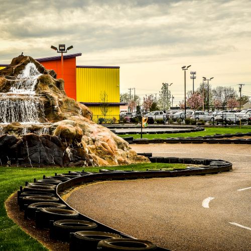 This image features a go-kart track with black tire barriers, a rock formation with a waterfall, green grass, and a colorful building in the background.