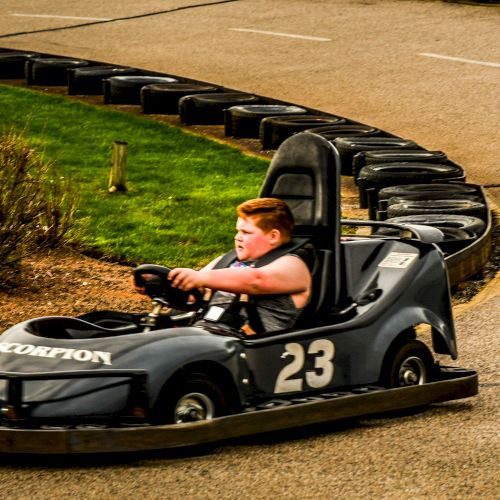 A person is driving a go-kart with the number 23 on a track bordered by tires and greenery, wearing a gray shirt.