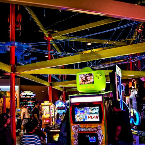 This image shows a vibrant indoor arcade with various game machines, colorful lights, and people enjoying different activities in the background.