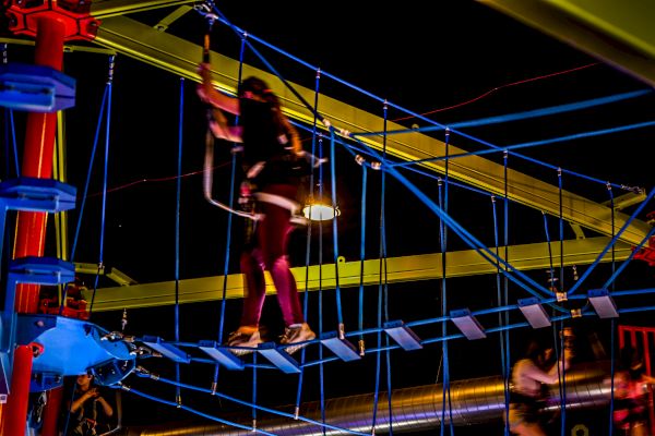 A person navigates through a brightly lit, indoor high ropes course at night. They are strapped in and walking across a suspended bridge.