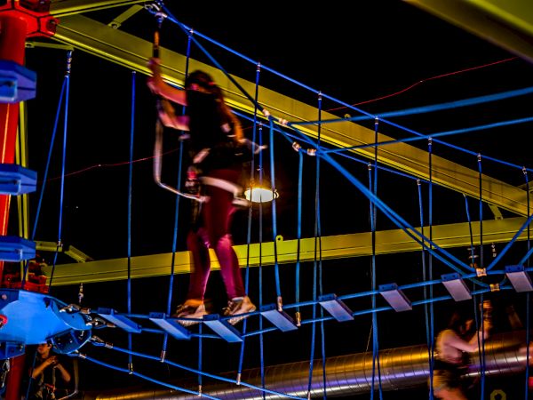 A person navigates through a brightly lit, indoor high ropes course at night. They are strapped in and walking across a suspended bridge.