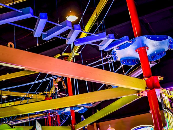 The image shows an indoor ropes course with colorful beams, ropes, and platforms, part of an adventure or amusement area.
