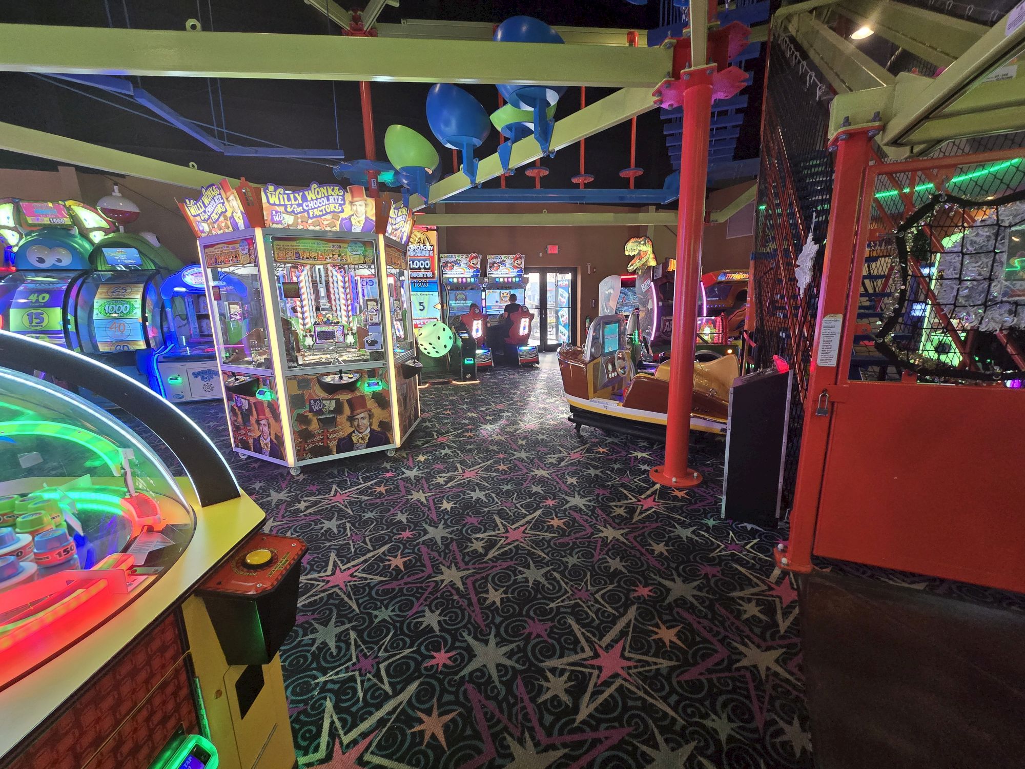 An arcade full of colorful claw machines, prize games, and neon lights under a starry carpet pattern, with balloons and bright, upbeat decor.
